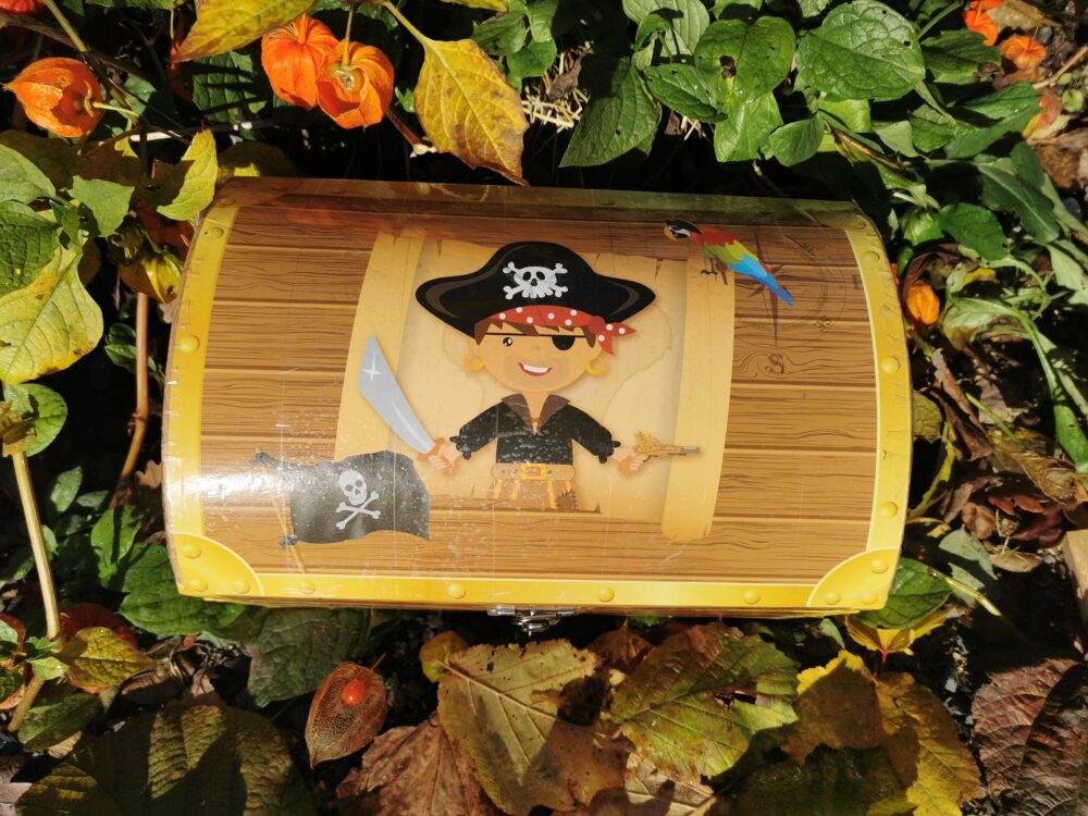 Eine Schatzkiste mit einem Piratenmotiv, das einen Piraten mit Schwert und Pistole zeigt. Die Kiste hat eine Holzoptik und ist mit einem Totenkopf-Symbol verziert.