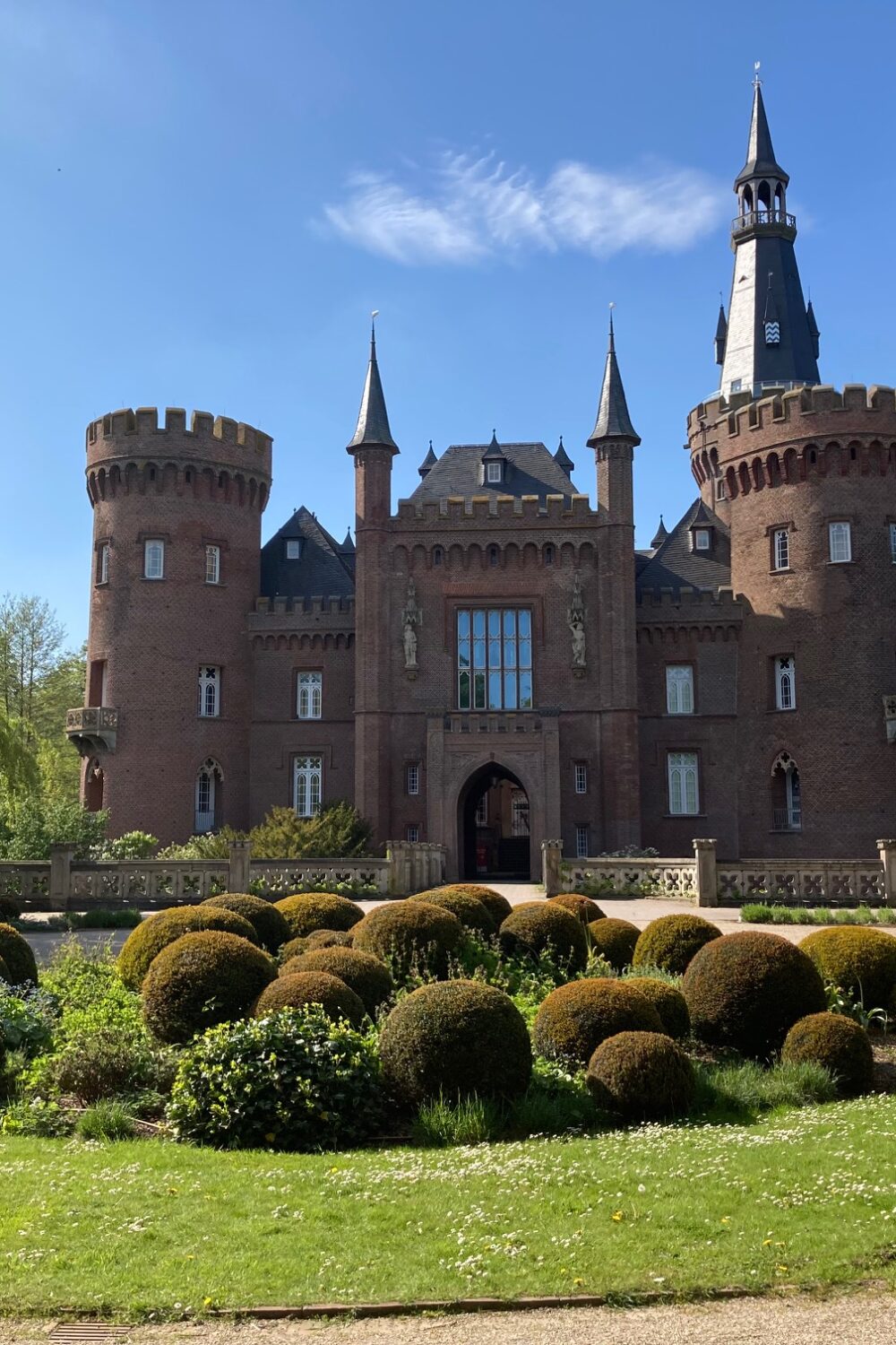 Rotes Backsteinschloss Moyland mit zwei runden Türmen, spitzen Dächern und einem Torbogen, davor ein runder Garten mit kugelförmigen Hecken.