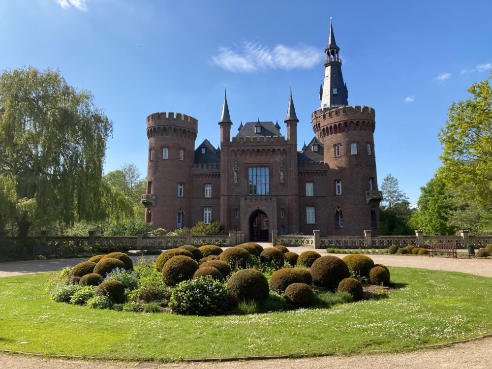 Rotes Backsteinschloss Moyland mit zwei runden Türmen, spitzen Dächern und einem Torbogen, davor ein runder Garten mit kugelförmigen Hecken.