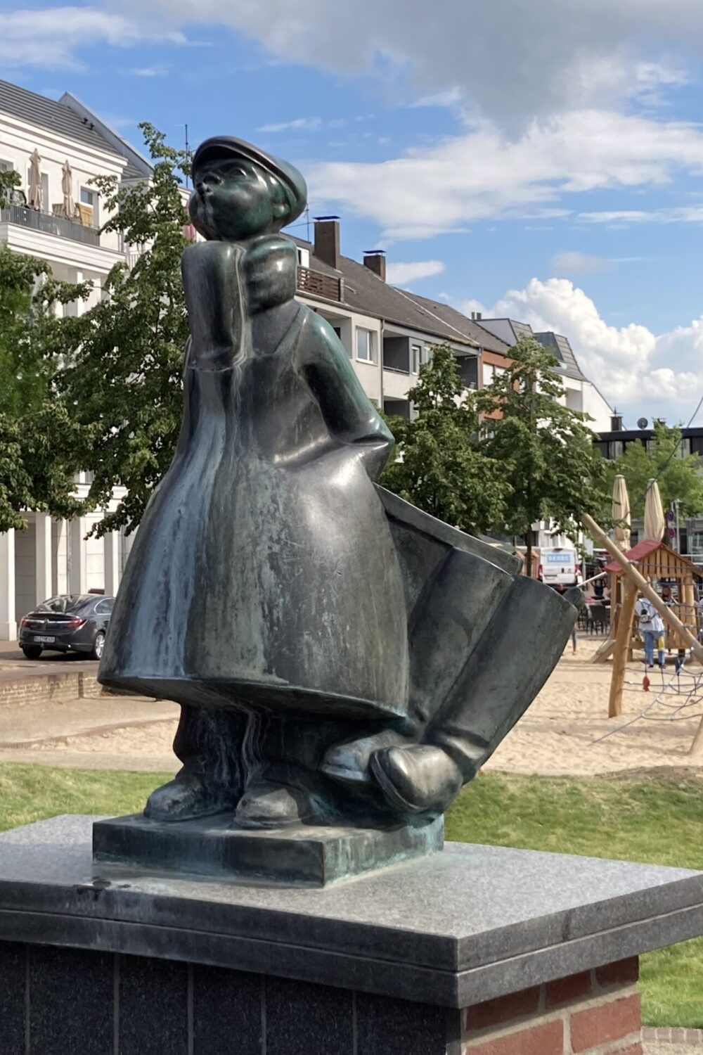 Bronzeskulptur des Schüsterken, einer Figur in traditioneller Kleidung mit einem großen Hut und einem Umhang, steht auf einem Sockel. Im Hintergrund sind Spielgeräte und Wohngebäude sichtbar.