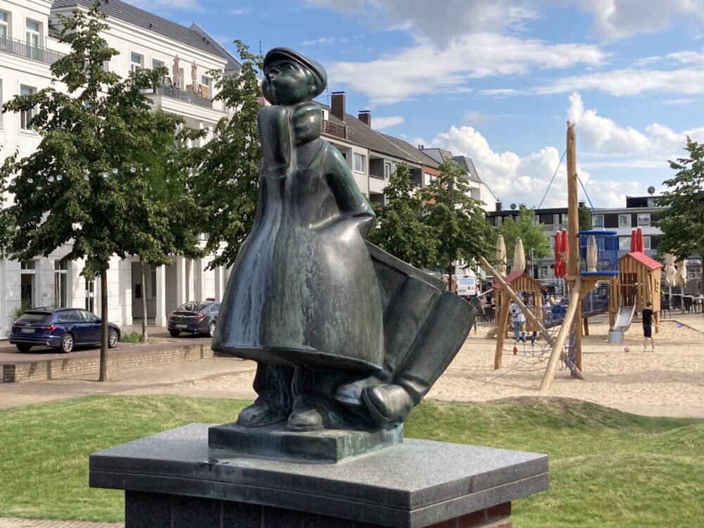 Bronzeskulptur des Schüsterken, einer Figur in traditioneller Kleidung mit einem großen Hut und einem Umhang, steht auf einem Sockel. Im Hintergrund sind Spielgeräte und Wohngebäude sichtbar.