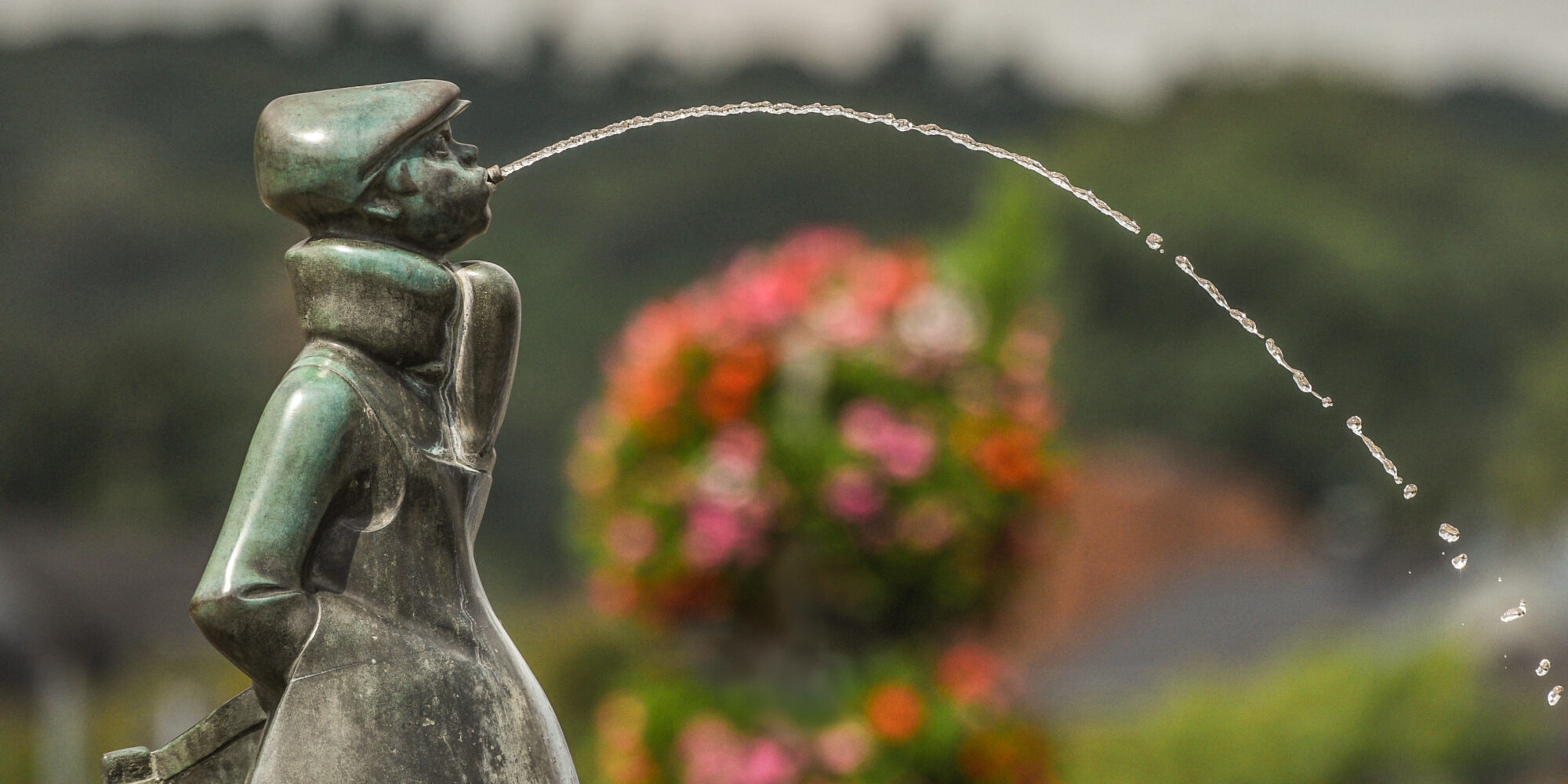 Bronze-Skulptur eines Jungen mit einer Mütze, der Wasser aus dem Mund spritzt. Im Hintergrund blühende Pflanzen und unscharfe Landschaft.