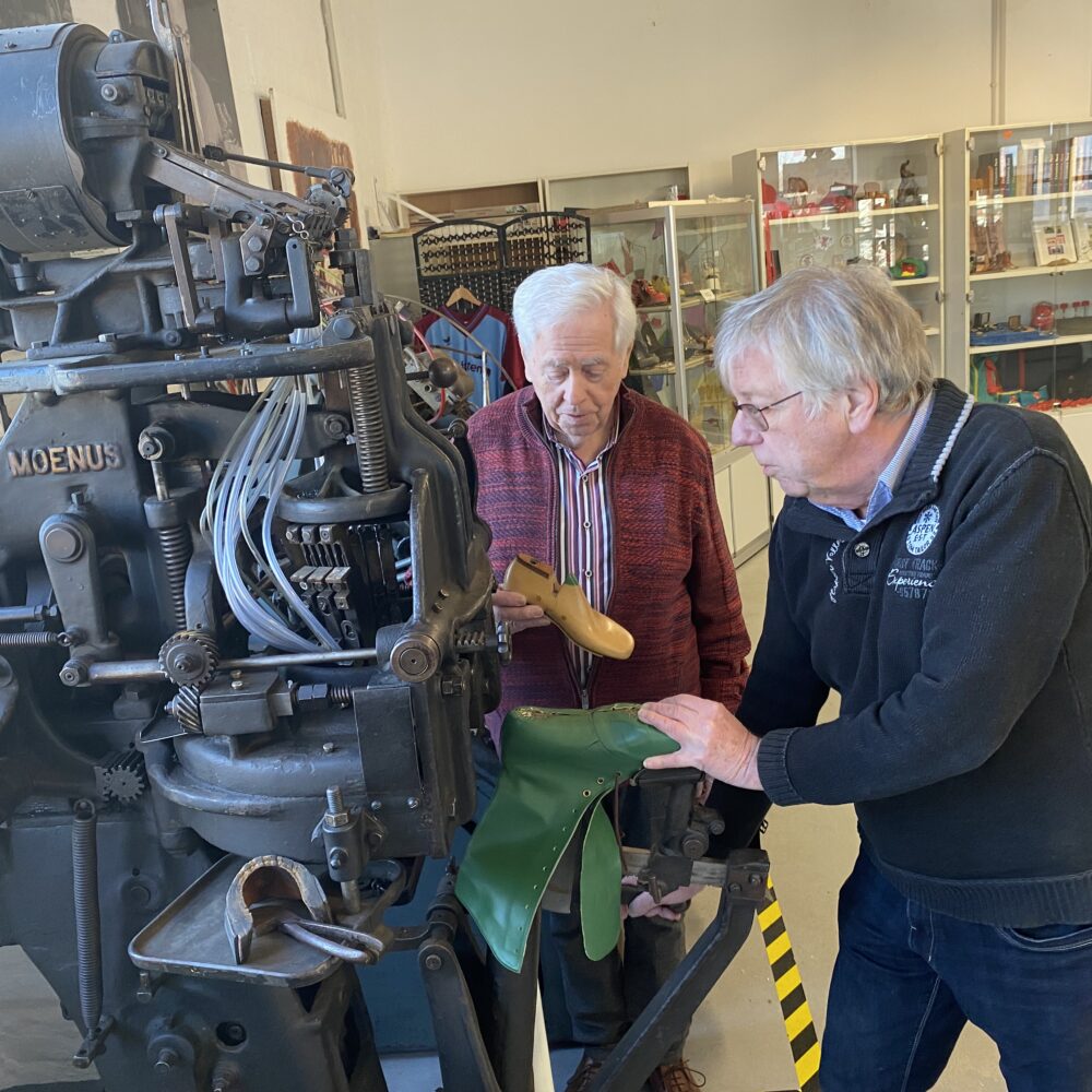 Zwei Ehrenamtliche des Klever Schuhmuseums stehen neben einer großen, schwarzen Schuhmachermaschine mit der Aufschrift 'MOENUS' im Schuhmuseum mit Vitrinen voller Schuhwerkzeuge.