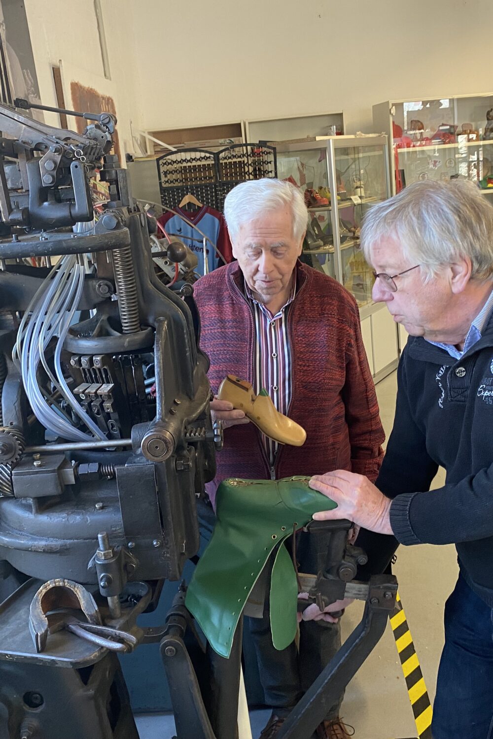 Zwei Ehrenamtliche des Klever Schuhmuseums stehen neben einer großen, schwarzen Schuhmachermaschine mit der Aufschrift 'MOENUS' im Schuhmuseum mit Vitrinen voller Schuhwerkzeuge.