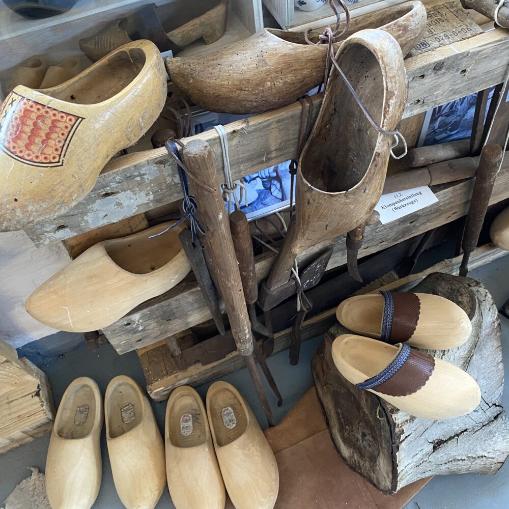 Verschiedene Holzschuhe im Klever Schuhmuseum, darunter bemalte und naturbelassene Modelle, einige hängen an einem Holzgestell, andere liegen auf Holzstücken.