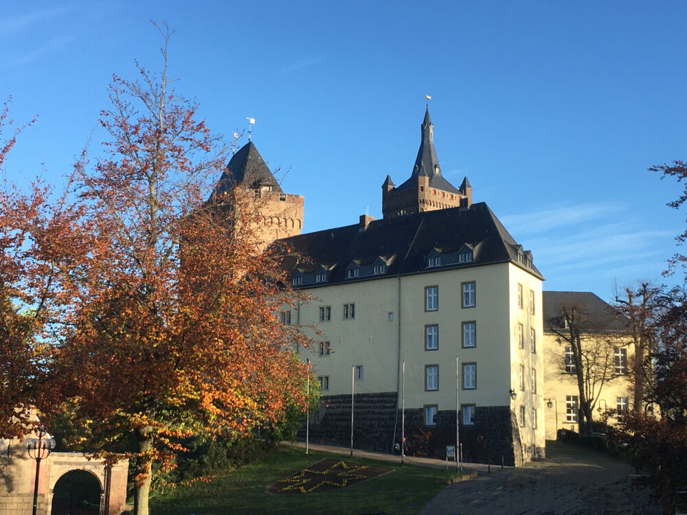 Die Schwanenburg mit zwei Türmen und einem gelben Gebäude im Vordergrund. Bunte Herbstblätter an einem Baum im Vordergrund.