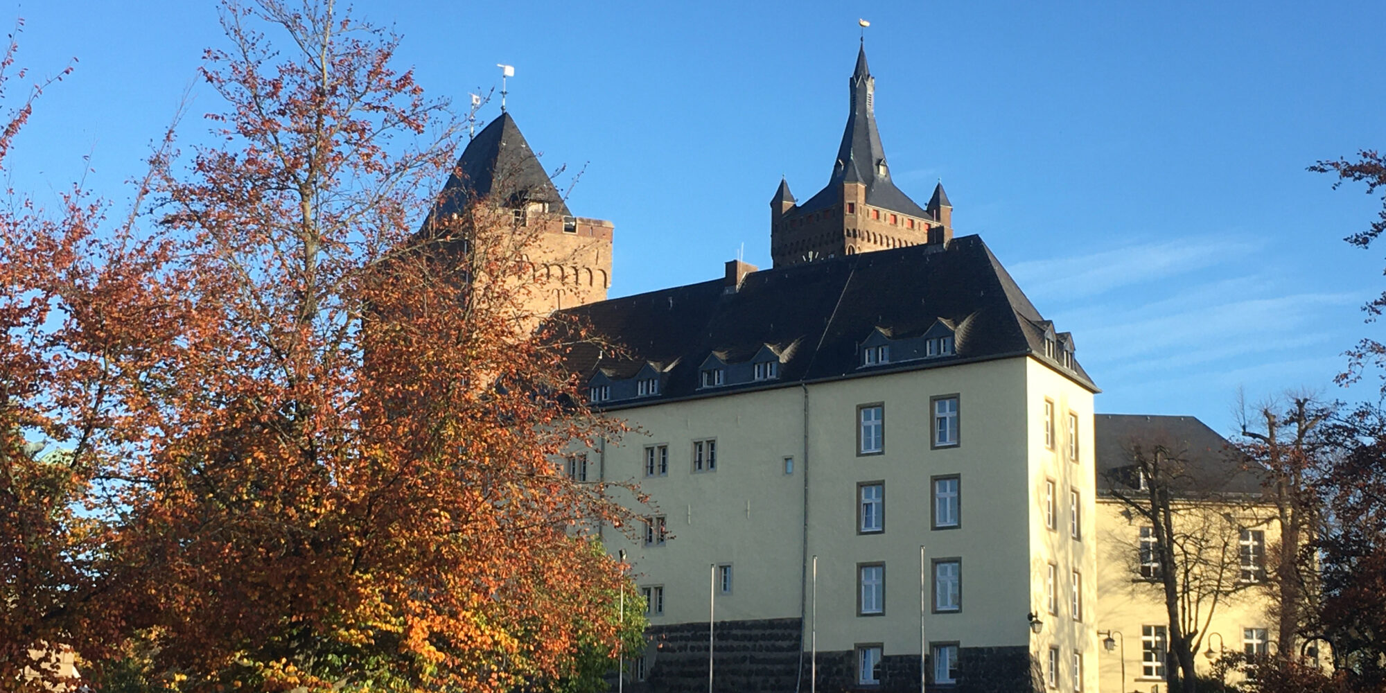 Die Schwanenburg mit zwei Türmen und einem gelben Gebäude im Vordergrund. Bunte Herbstblätter an einem Baum im Vordergrund.