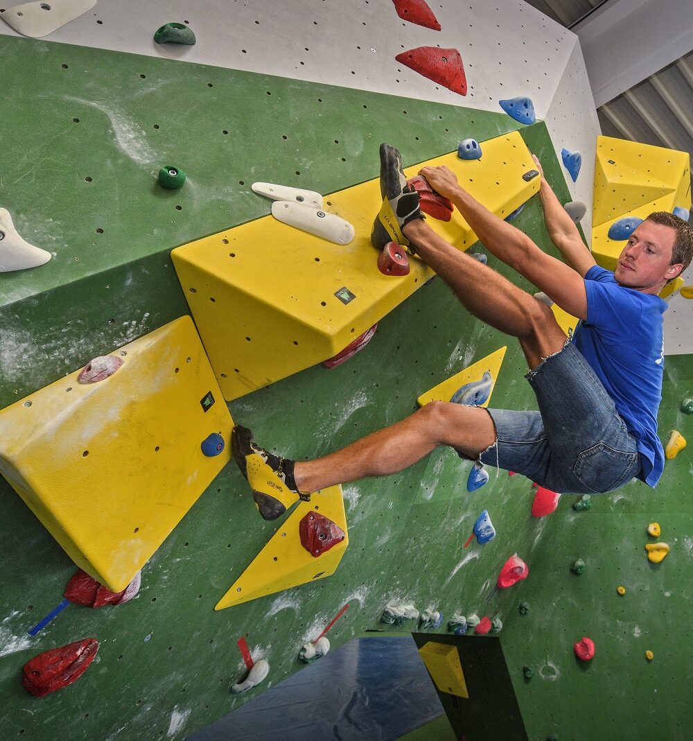 Junger Mann im blauen T-Shirt und Shorts klettert an einer grünen Kletterwand in der Kliff-Boulderhalle mit gelben und roten Griffen. Er hat einen Fuß auf einem gelben Griff und zieht sich mit den Händen hoch.