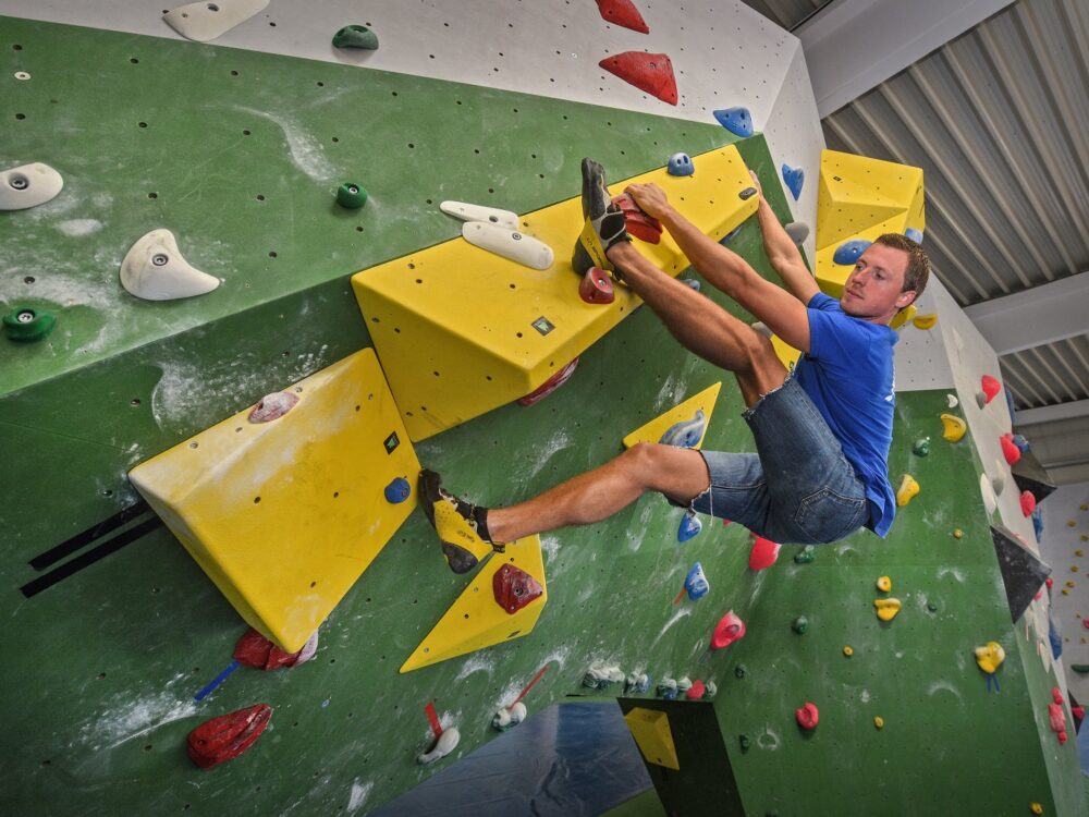 Junger Mann im blauen T-Shirt und Shorts klettert an einer grünen Kletterwand in der Kliff-Boulderhalle mit gelben und roten Griffen. Er hat einen Fuß auf einem gelben Griff und zieht sich mit den Händen hoch.