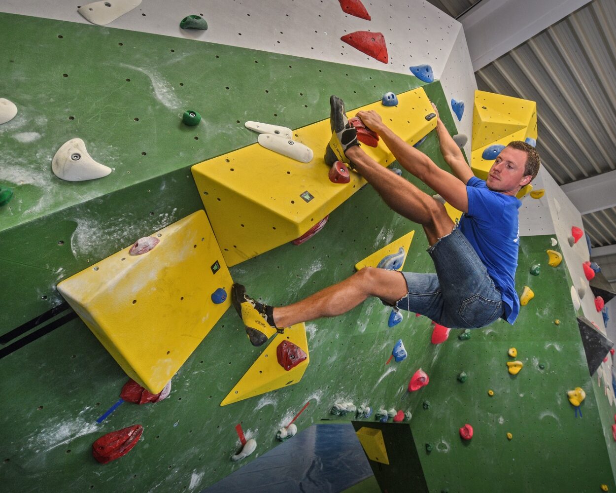 Junger Mann im blauen T-Shirt und Shorts klettert an einer grünen Kletterwand in der Kliff-Boulderhalle mit gelben und roten Griffen. Er hat einen Fuß auf einem gelben Griff und zieht sich mit den Händen hoch.