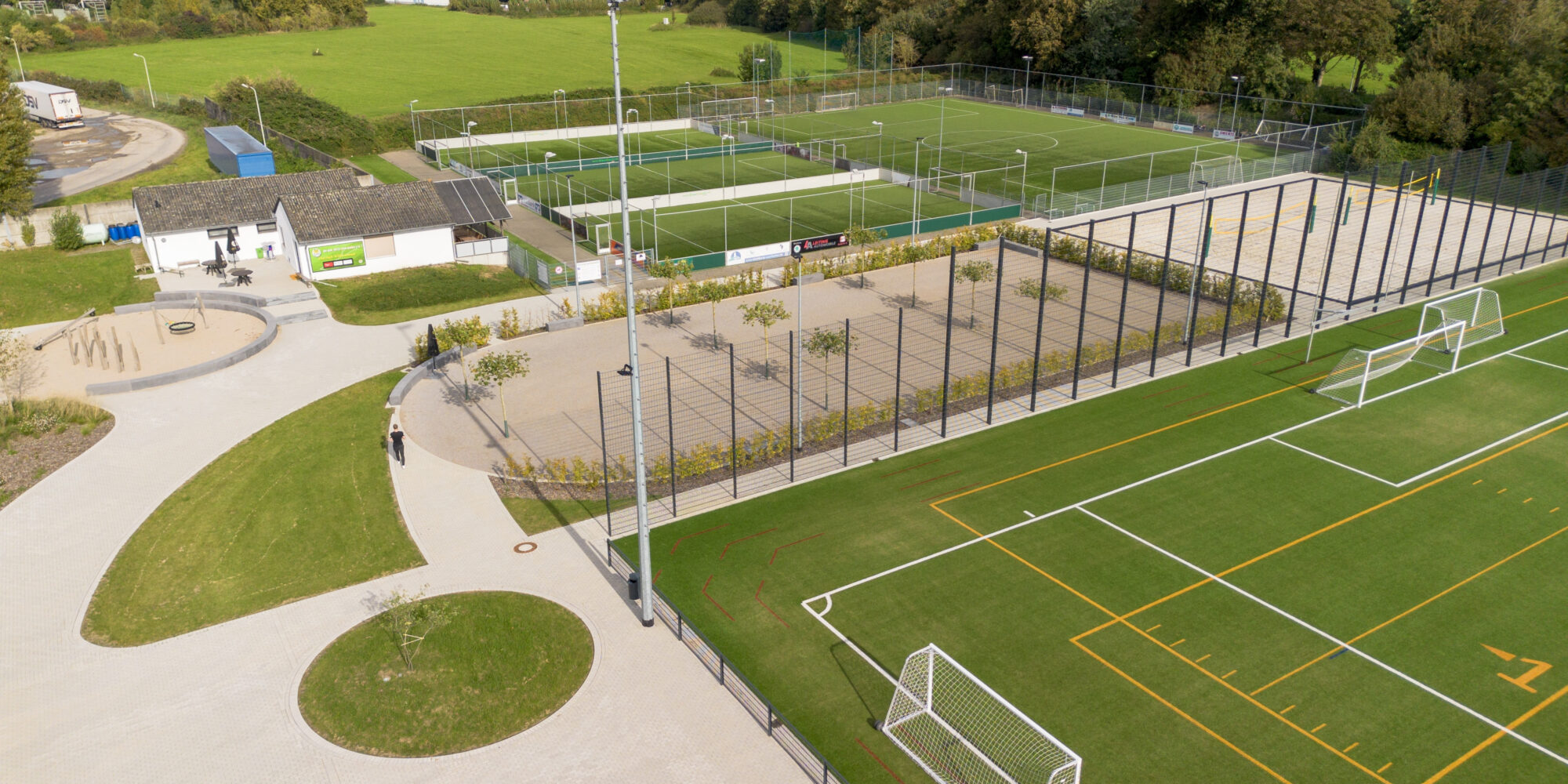 Luftaufnahme der Sportanlage Kellen mit mehreren Fußballfeldern, einem Parkplatz und einem angrenzenden Grünbereich. Im Hintergrund sind in der Erlebnisregion Kleves Bäume und ein Wohngebiet sichtbar.