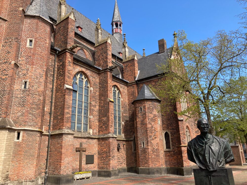Die Stiftskirche in Kleve, aus rotem Backstein mit mehreren hohen Fenstern und einem spitzen Turm. Im Vordergrund steht eine Statue.