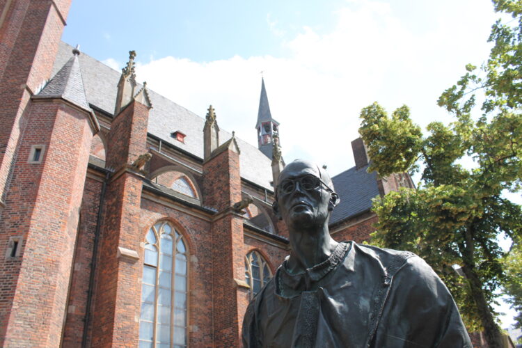 Bronzestatue von Karl Leisner, stehend im Vordergrund. Im Hintergrund die Stiftskirche mit roten Ziegelsteinen und einem hohen Kirchturm.