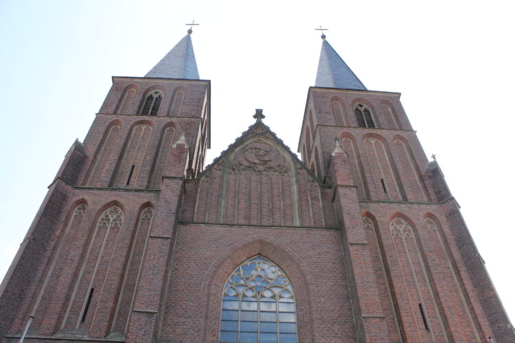 Fassade der Stiftskirche mit zwei hohen, spitzen Türmen, Ziegelsteinmauerwerk und einem großen, gotischen Fenster in der Mitte.