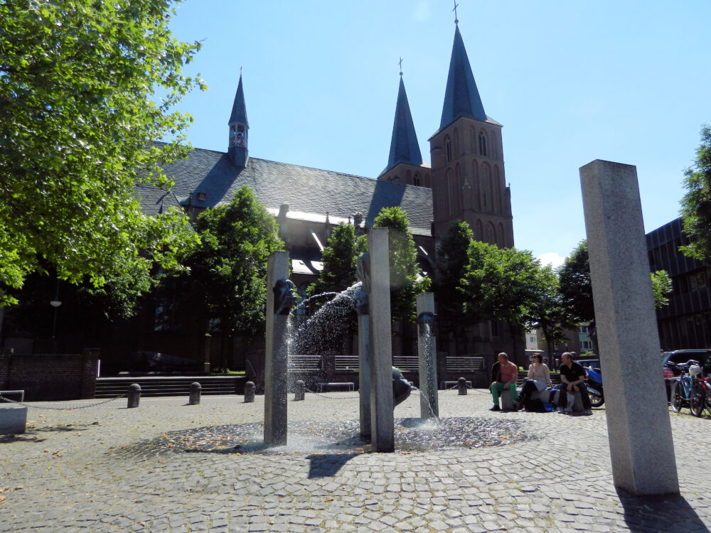 Stiftskirche mit zwei spitzen Türmen im Hintergrund, Narrenbrunnen im Vordergrund mit Wasserfontäne, umgeben von Bäumen und Sitzenden.
