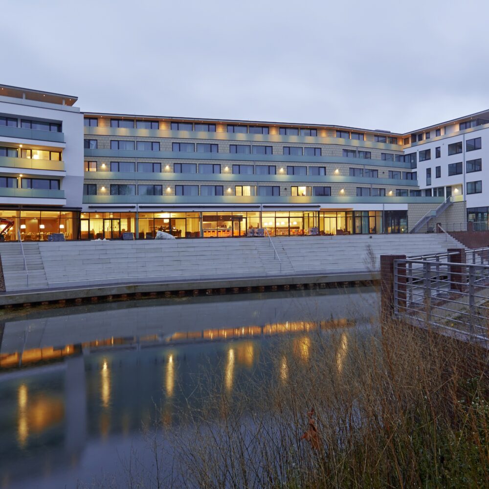 Modernes mehrstöckiges Gebäude des elaya hotel kleve mit großen Fenstern und beleuchteten Innenräumen am Wasserufer bei Dämmerung