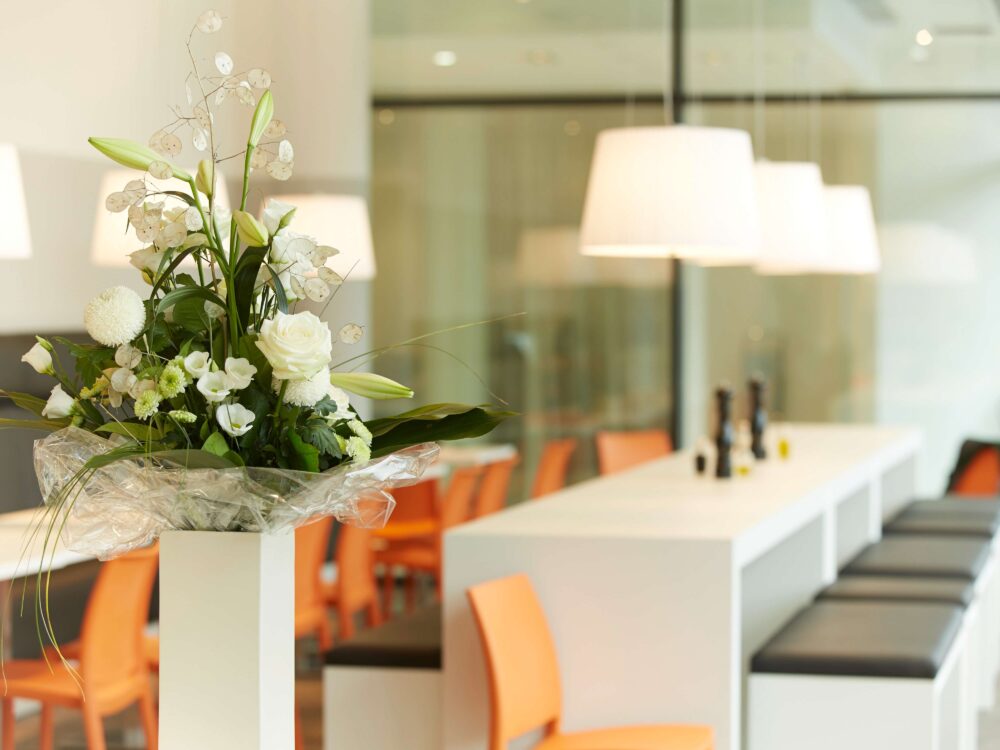 Weiß-grüner Blumenstrauß in Vase vor modernem Raum mit orangen Stühlen und weißen Lampen im elaya hotel kleve