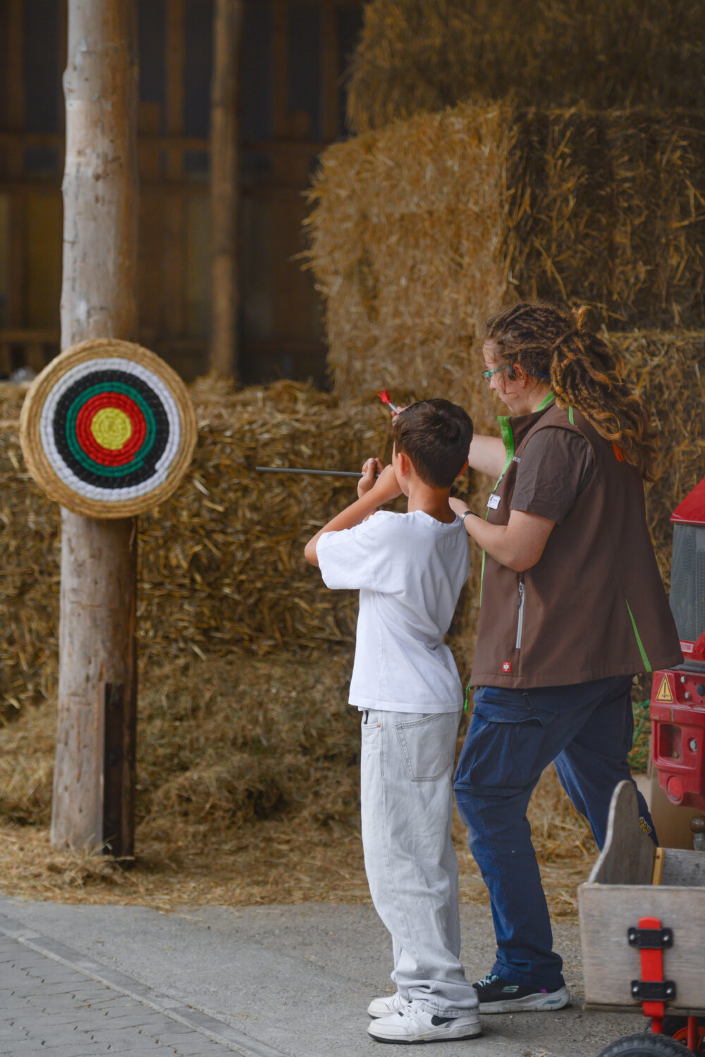 Ein Kind in einem weißen T-Shirt und eine Person mit langen, geflochtenen Haaren stehen vor einer Zielscheibe aus Stroh. Das Kind hält einen Pfeil, während die andere Person ihm assistiert.