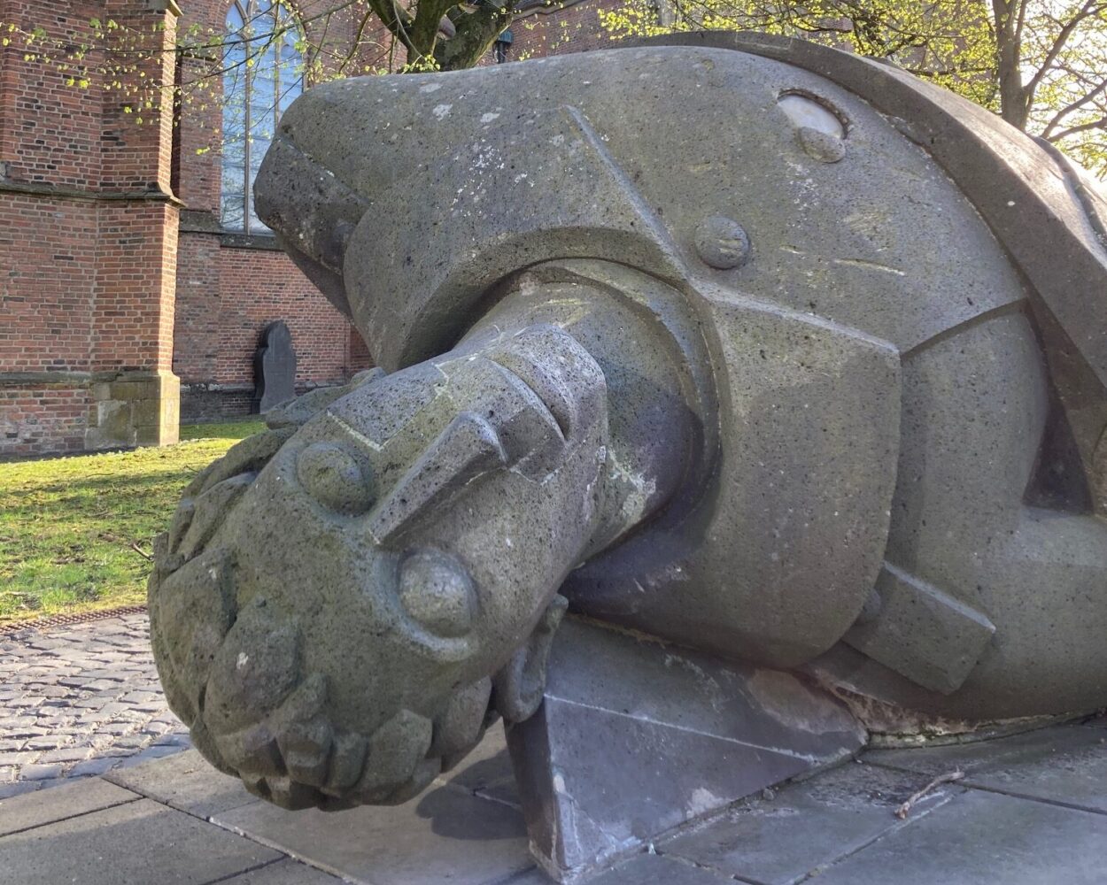 Der Tote Krieger: Skulptur eines toten Kriegers, liegend auf einem Sockel, umgeben von Bäumen und einer Kirche im Hintergrund. Der Krieger trägt eine Rüstung und hat einen ernsten Gesichtsausdruck.