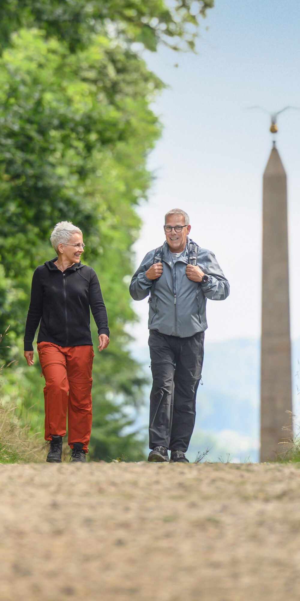 Zwei Personen gehen auf einem Kiesweg durch eine grüne Landschaft. Im Hintergrund steht ein hoher Obelisk.