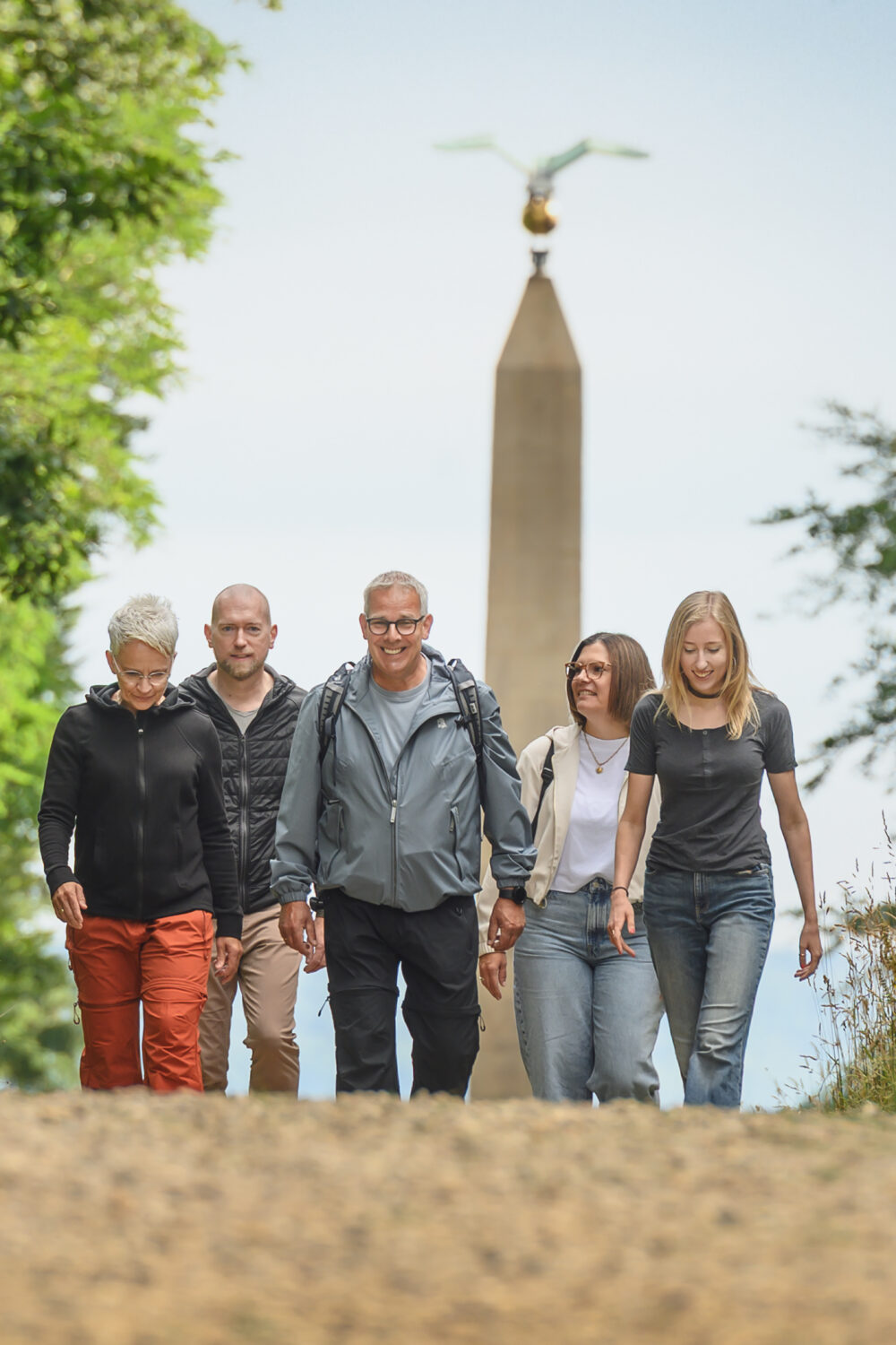 Fünf Personen gehen auf einem Kiesweg, umgeben von Bäumen und hohem Gras. Im Hintergrund steht ein Obelisk mit einer Statue oben.
