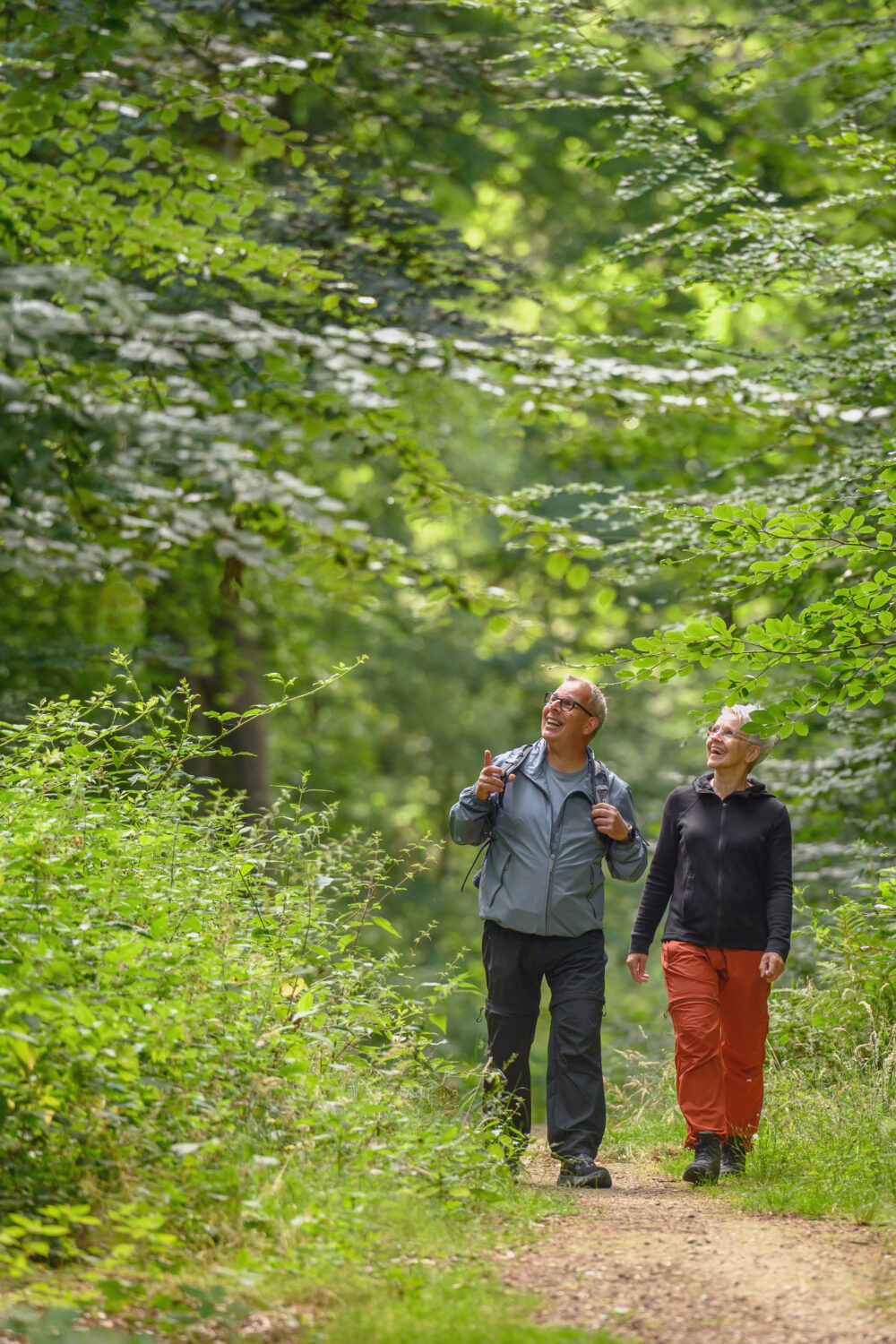 Zwei Personen wandern auf einem Waldweg im Reichswald, umgeben von Bäumen und Sträuchern. Eine Person trägt eine graue Jacke und die andere eine orange Hose.