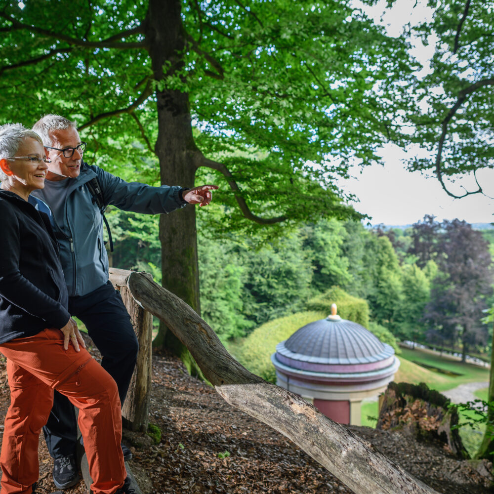 Zwei Personen stehen auf einem Waldweg neben einem Baumstamm, umgeben von grünen Bäumen, mit Blick auf ein rundes Gebäude mit Kuppeldach im Tal.
