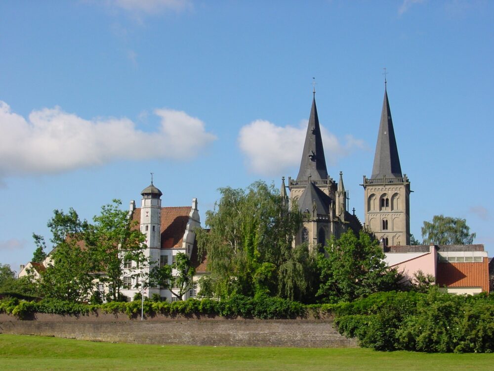 Blick auf die Türme einer gotischen Kirche hinter Bäumen und historischen Gebäuden unter blauem Himmel mit Wolken in Xanten.