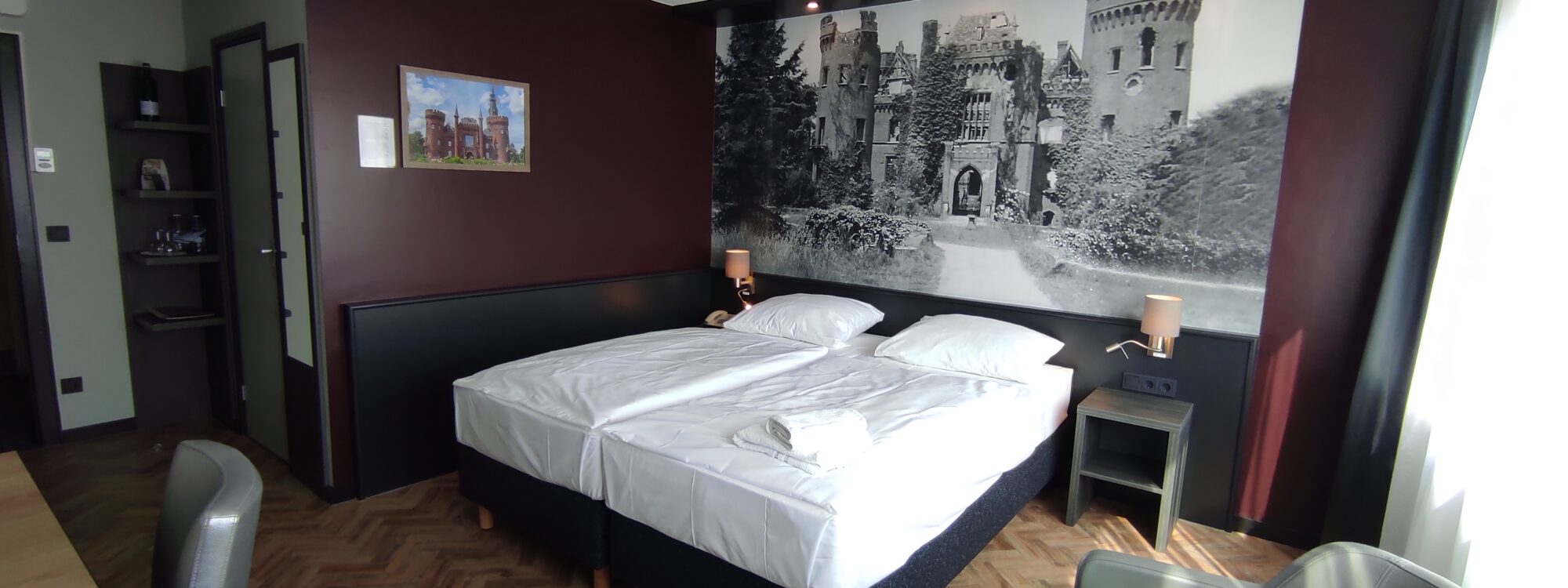 Unterkunft in Kleve. Hotelzimmer mit zwei Einzelbetten, Nachttischen mit Lampen, großem Schwarz-Weiß-Bild einer Burg an der Wand und einem runden Tisch mit zwei Stühlen
