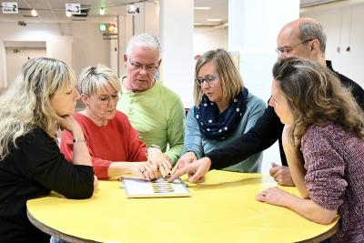 Indoor-Aktivitäten für Gruppen. Sechs Personen stehen um einen runden Tisch und zeigen auf ein Blatt Papier. Zwei Frauen tragen rote und graue Oberteile, eine Frau hat einen blauen Schal, und die anderen tragen Pullover.