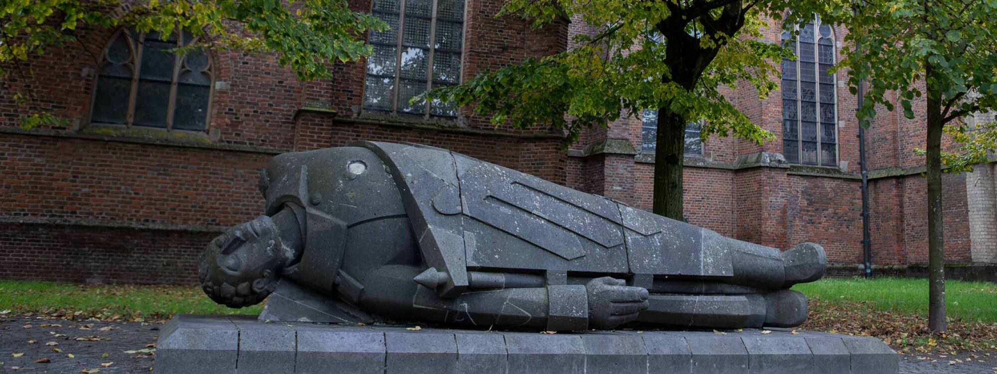 Der Tote Krieger: Skulptur eines toten Kriegers, liegend auf einem Sockel, umgeben von Bäumen und einer Kirche im Hintergrund. Der Krieger trägt eine Rüstung und hat einen ernsten Gesichtsausdruck.