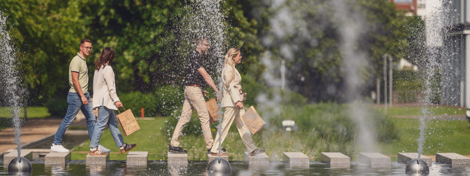 Eine Gruppe von jungen Menschen läuft mit Tüten an einem Wasserspiel vorbei