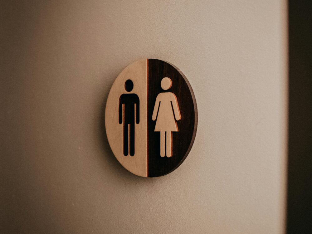 Rundes Schild mit stilisierten männlichen und weiblichen Figuren, halb hell, halb dunkel, an Wand montiert.