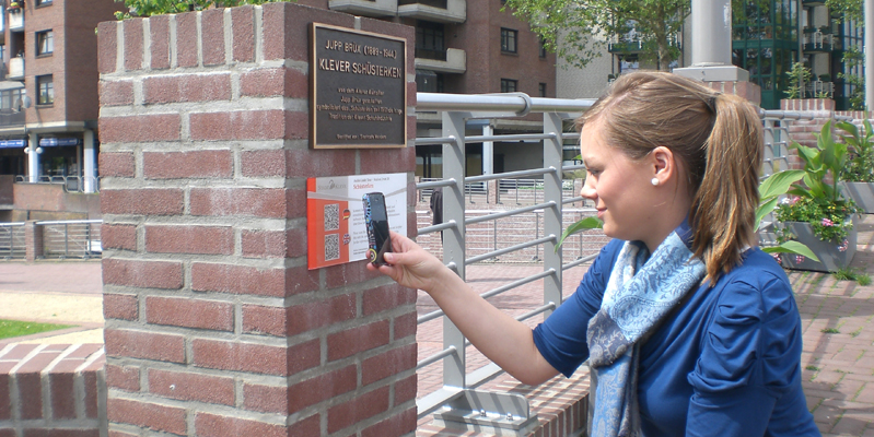 Frau mit blauem Tuch scannt QR-Code an einer Backsteinmauer mit Gedenktafel und Metallgeländer im Hintergrund.
