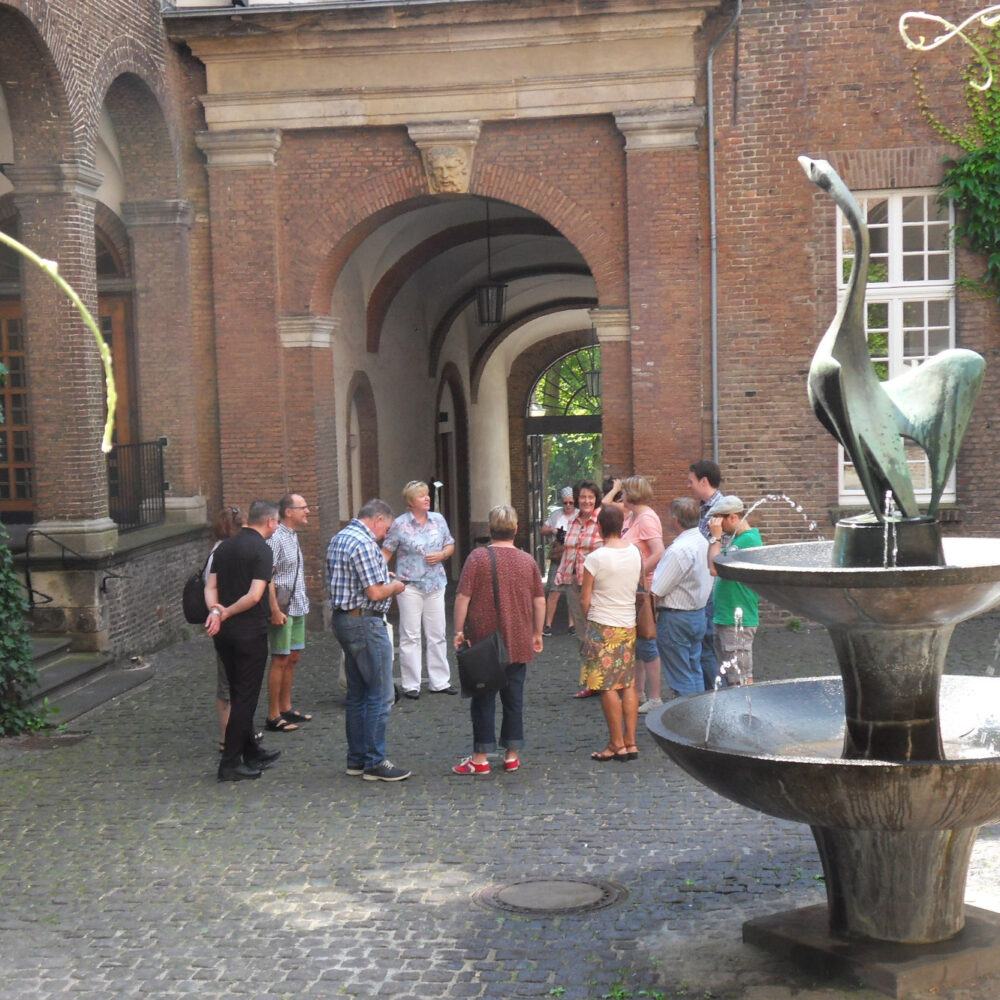 Gruppe von Menschen steht in einem Innenhof der Schwanenburg mit Kopfsteinpflaster und Backsteinbögen, rechts ein Brunnen mit Vogel-Skulptur und Wasserstrahlen