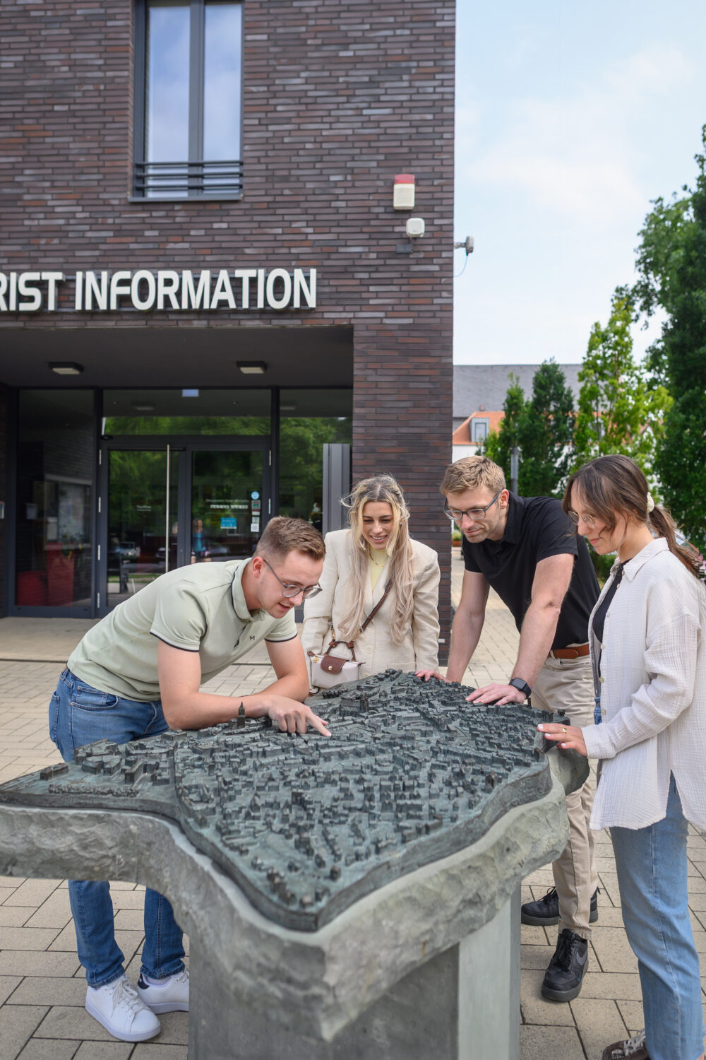 Vier Personen stehen um ein dreidimensionales Stadtmodell aus Stein von Kleve vor einem Gebäude mit der Aufschrift 'TOURIST INFORMATION'.