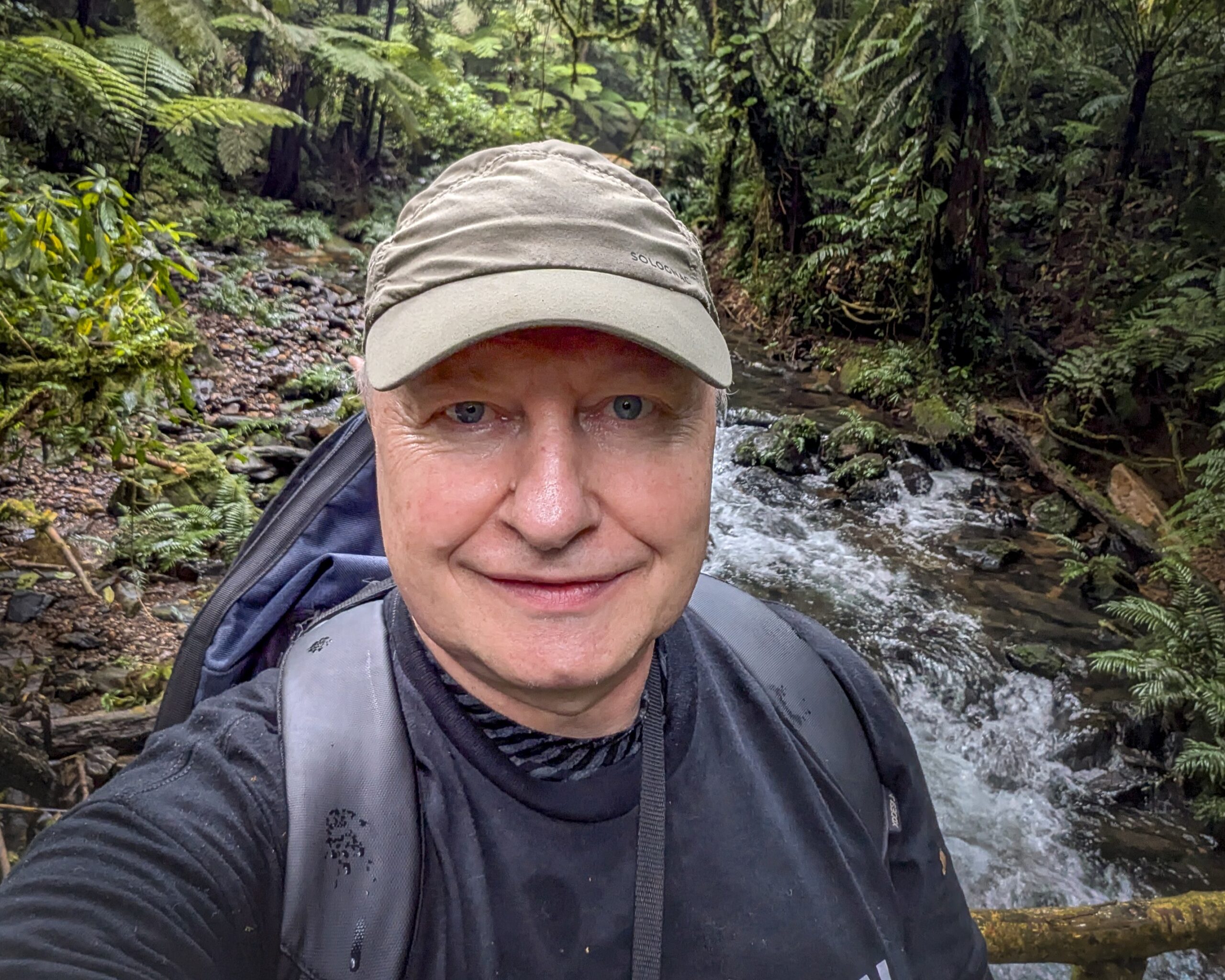 Professor Dr. Dirk Reiser mit hellgrauer Kappe, schwarzem T-Shirt und Rucksack vor einem Bach in einem dichten, grünen Wald.