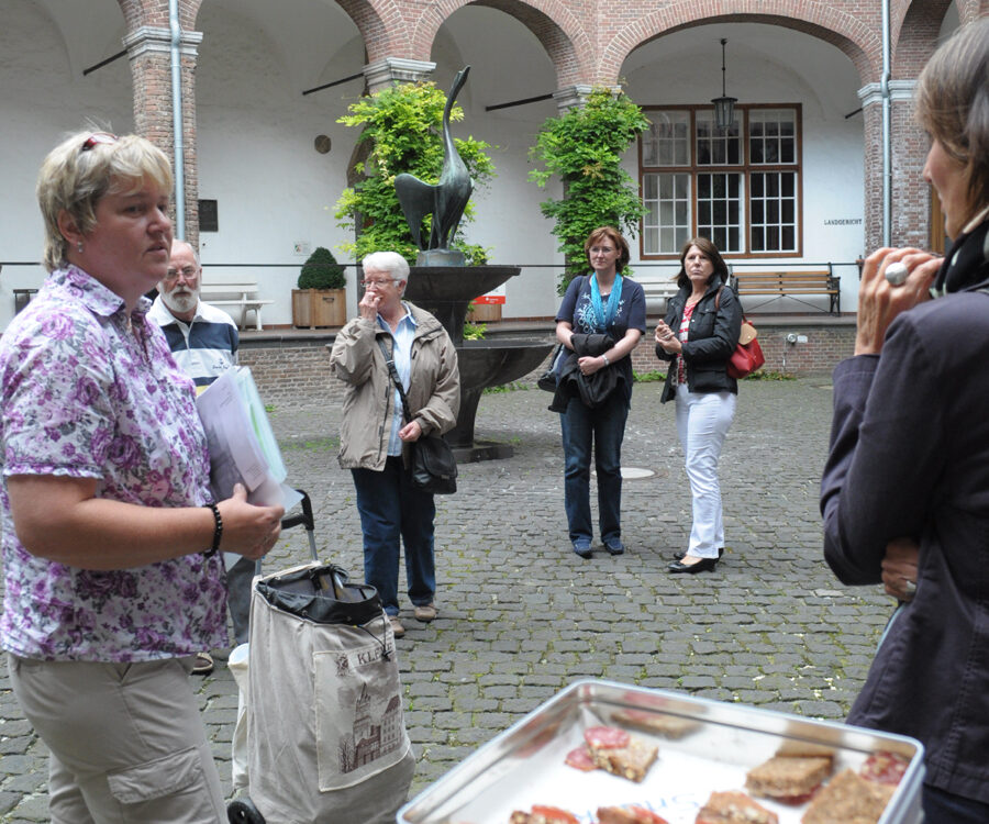 Mehrere Personen stehen vor einem Backsteingebäude, eine Person hält eine weiße Plastikbox mit kleinen Speisen darin.