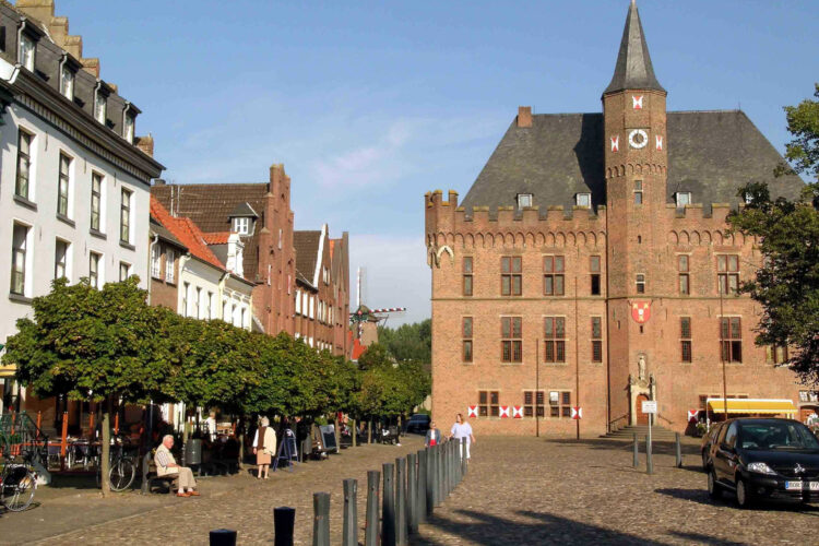 Historisches Backsteingebäude mit Turm und Zinnen am Marktplatz, links Baumreihe und Menschen auf Bänken und Gehweg.