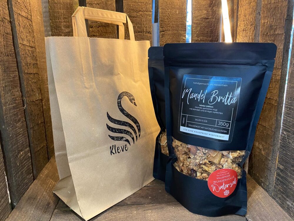 Das Bild zeigt mehrere schwarze Tüten mit dem Müsli Mandel Britta rechts und eine Einkaufstasche aus Papier mit Kleve Schwan links. Die Tüten stehen in einer Holzkiste.