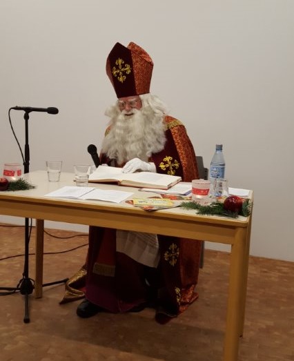 Nikolaus mit rotem Bischofsgewand und Mitra sitzt an einem Tisch mit offenem Buch, Mikrofon und Wasserflasche.