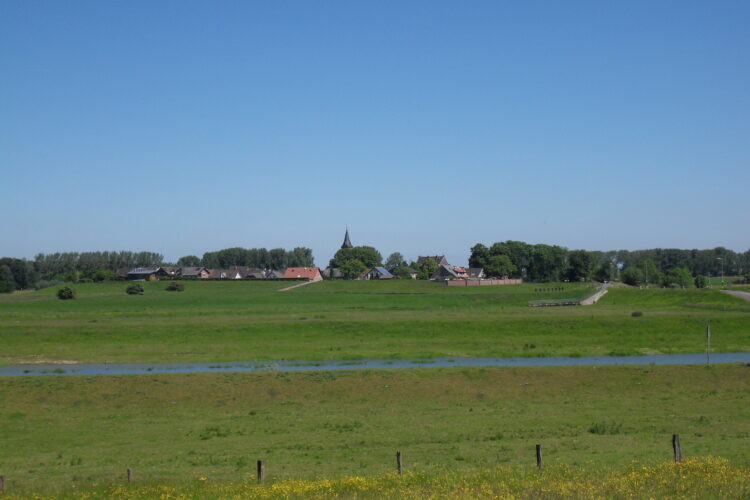 Weite grüne Wiesen mit einem schmalen Wasserlauf im Vordergrund und einem Dorf mit Kirchturm im Hintergrund unter blauem Himmel.
