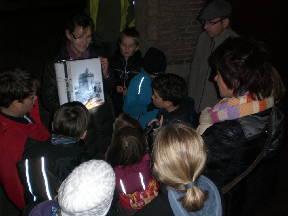 Gruppe von Kindern und Erwachsenen im Dunkeln, eine Person hält eine beleuchtete Mappe mit einem Bild, mehrere tragen reflektierende Kleidung oder Accessoires.