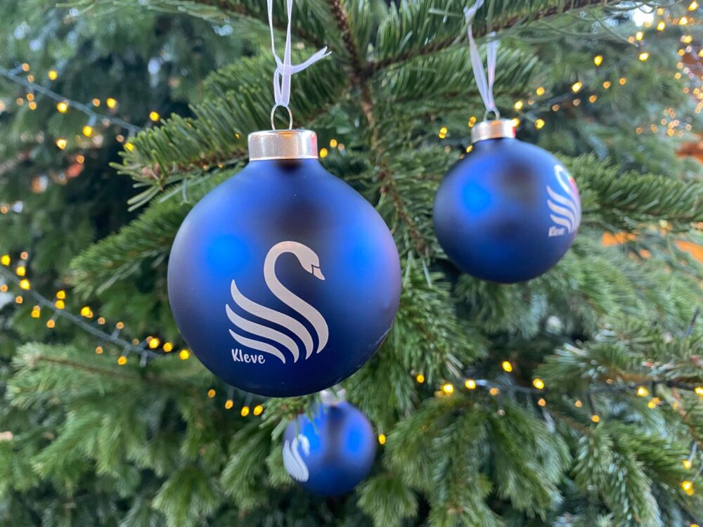 Drei blaue Weihnachtskugeln mit weißem Schwan-Logo und Schriftzug 'Kleve' hängen an einem Tannenzweig mit Lichterkette im Hintergrund.